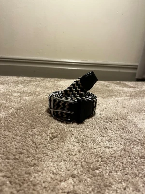 Flätat bälte från Ragazzo  - Ett stilrent flätat bälte i färgerna svart blått vitt och beige. 125cm lång. Hör av er vid frågor eller funderingar 