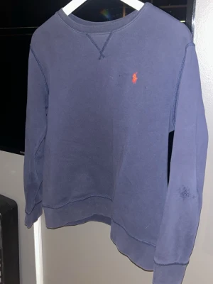 Blå sweatshirt från Ralph Lauren 160-168 cm - Mörkblå långärmad sweatshirt från Ralph Lauren med klassisk orange logga broderad på bröstet. Tröjan har rund hals och ribbade muddar vid ärmslut och nederkant. Perfekt för chill dagar och enkel att matcha med jeans eller joggers.