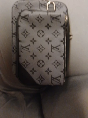 Väska  i grått - Snygg datorväska från  i grått med klassiskt monogrammönster. Väskan har en rektangulär form, dragkedja och detaljer i svart skinn. Perfekt för dig som vill ha en stilren och lyxig accessoar till din laptop.