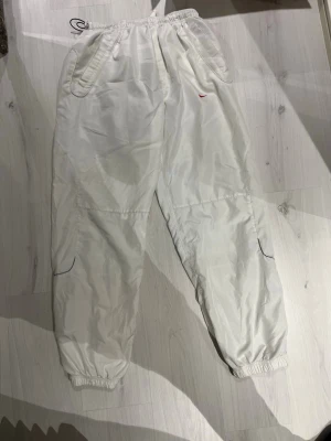 Vita Nike träningsbyxormed röd logo - Säljer ett par vita träningsbyxor från Nike med röd swoosh på framsidan. Byxorna har elastisk midja och benslut, samt praktiska fickor på sidorna. Materialet är syntet.