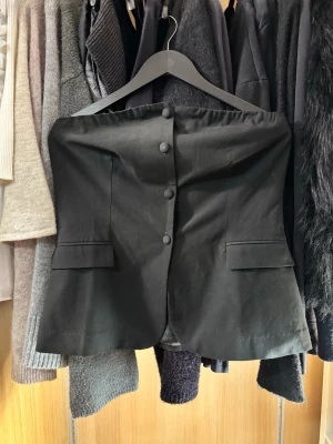 Festtopp  - Säljer en svart tubtopp med knappar framtill och snygga fickdetaljer. Toppen har en bandeauformad överdel och är figurnära med lätt utsvängd nederkant. Storlek M från H&M.