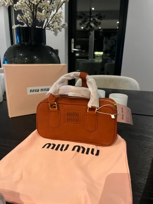 Brun handväska från Miu Miu mocka - Säljer en snygg brun handväska från Miu Miu i mjuk mocka. Väskan har guldfärgade metalldetaljer, dubbla handtag och dragkedja upptill. Klassisk rektangulär form med Miu Miu-logga framtill. Perfekt accessoar för att lyfta din outfit.