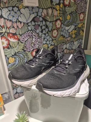 Svarta Hoka One One Arahi 5 sneakers - Säljer ett par svarta Hoka One One Arahi 5 sneakers med vit och grå sula. Skorna har ventilerande mesh-ovandel, snörning och reflekterande detaljer på sidorna. Perfekta för dig som gillar att springa eller träna och vill ha riktigt skön dämpning.