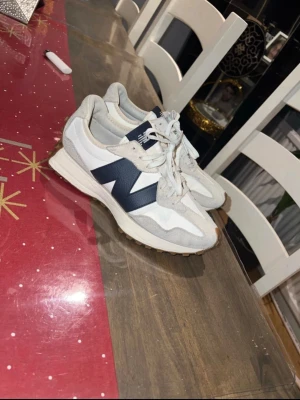 New Balance vita och marinblå sneakers - Säljer ett par snygga New Balance sneakers i vitt och marinblått med grå mockadetaljer. Skorna har snörning, rund tå och en platt sula med bruna gummidetaljer på hälen. Perfekt för dig som gillar stilrena och sportiga sneakers.