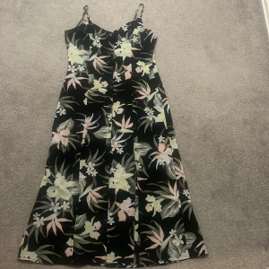 Svart långklänning med blommigt mönster - Säljer en svart långklänning med tunna axelband och knappar framtill. Klänningen har ett färgstarkt blommigt mönster i ljusa toner av grönt, gult och rosa. Materialet känns mjukt och har ett lätt fall, perfekt för varma dagar.