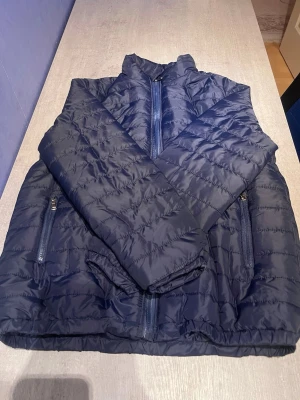 Mörkblå pufferjacka med dragkedja - Säljer en mörkblå pufferjacka med hög krage och två sidofickor med dragkedja. Jackan är quiltad och har långa ärmar samt elastisk nederkant. Perfekt för kyliga dagar och riktigt lätt att bära. Passar dig som gillar enkel och stilren look.