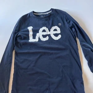 Mörkblå långärmad t-shirt från Lee - Snygg mörkblå långärmad t-shirt från Lee med stort vitt logotryck över bröstet. Klassisk rund halsringning och mjukt bomullsmaterial som känns skönt mot huden. Perfekt för en avslappnad och sopad look.