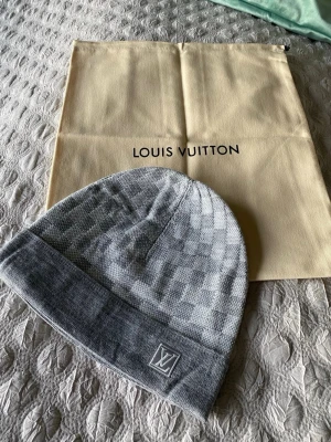 Grå LV Mössa - Säljer en snygg grå mössa från Louis Vuitton med klassiskt rutigt mönster och LV-logga framtill. Mössan är stickad och har en bred uppvikt kant. Perfekt för dig som vill ha en lyxig accessoar till vintern.