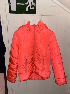 Korall färgad dunjacka från Lindex - Säljer en snygg korall färgad dunjacka från Lindex med huva och två fickor med dragkedja. Jackan har en reflekterande detalj baktill. Perfekt för dig som gillar färg och vill sticka ut lite extra under höst och vinter.
