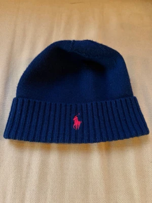 Mörkblå ullmössa från Polo Ralph Lauren - Snygg mörkblå mössa från Polo Ralph Lauren i 100% ull. Mössan har ribbad kant och en rosa broderad logga framtill. Perfekt för att hålla värmen och samtidigt se stilren ut.