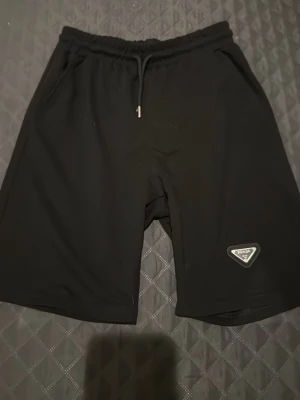 Svarta shorts från Prada med snörning - Säljer ett par svarta shorts från Prada med elastisk midja och snörning framtill. De har en stilren design med Prada-logga på vänster ben och är tillverkade i ett mjukt syntetmaterial. Perfekta för chill dagar och streetwear.