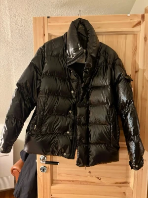 Svart pufferjacka från Moncler - Säljer en svart, glansig pufferjacka från Moncler med broderad logga på ärmen. Jackan har hög krage, dragkedja och knappar framtill samt två sidofickor. Materialet är syntet och dun, perfekt för kalla dagar. Stilsäker och riktigt fet jacka för dig som vill sticka ut.