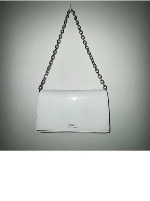 Vit axelväska från Polo Ralph Lauren - Vit axelväska från Polo Ralph Lauren med krokodilmönster och silvrig kedjerem. Sömmarna runt om väskan är svarta vilket gör den mer unik. Kan användas med långt band, runt om kroppen. Även med nedkortat band då den passar perfekt som axelväska. Samt som kuvertväska. 
