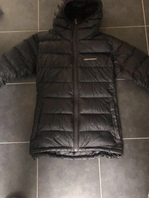 Svart Peak Performance dunjacka - Svart dunjacka från Peak Performance med huva och dragkedja framtill. Jackan har ett quiltat mönster och logga på bröstet. Perfekt för kalla dagar och har en snygg, normal passform. Materialet är polyester och dun, vilket gör den både lätt och väldigt varm. Kan gå ner i pris vid snabb affär!!