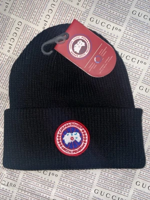 Svart Canada Goose mössa med patch - Svart ribbstickad mössa från Canada Goose med klassisk rund patch framtill i rött, vitt och blått. Mössan har uppvikt kant och är tillverkad i polyester och bomull. Perfekt för kalla dagar och har en snygg streetstil.