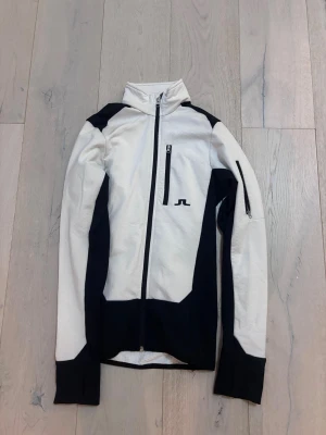 Vit och svart vindjacka från J.Lindeberg S/XS - Snygg vindjacka från J.Lindeberg i vit och svart med hög krage och hel dragkedja framtill. Jackan har en bröstficka med dragkedja och logga på bröstet samt på kragen. Långärmad och sportig design, perfekt för aktiva dagar.