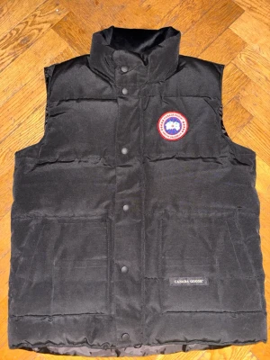 Svart dunväst från Canada Goose - Svart dunväst från Canada Goose med hög krage och tryckknappar framtill. Klassisk broderad logotyp på bröstet och två stora fickor framtill. Västen är quiltad och har en clean, stilren look som funkar till många outfits.( Priset kan diskuteras vid SNABB affär)😁