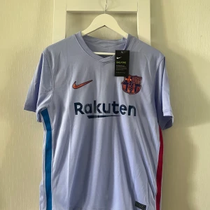 FC Barcelona bortatröja Nike S - Snygg FC Barcelona bortatröja från Nike i ljusblått med klubbmärke och Rakuten-tryck på bröstet. Tröjan har korta ärmar, rund hals och färgdetaljer i blått och rött på sidorna. Tillverkad i lätt och ventilerande polyester, perfekt för fotboll eller supporterliv.