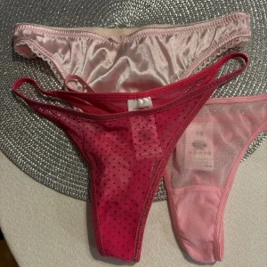 Rosa och röda stringtrosor XS - Tre par stringtrosor i olika nyanser av rosa . Ett par är ljusrosa i glansigt satinmaterial, ett par är mörkrosa med små prickar och mesh, och det tredje paret är ljusrosa mesh. Perfekt för dig som gillar snygga och bekväma underkläder.