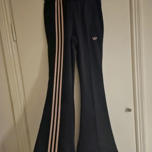 Svarta bootcut byxor från Adidas XS - Svarta Adidas byxor i bootcut-modell med ljusbeige ränder längs benen och klassisk Adidas-logga på höften. Byxorna har dragkedjeförsedda fickor och är gjorda i mjukt syntetmaterial. Perfekt för dig som gillar sportig och retro stil.