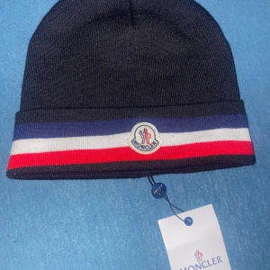 Moncler mössa - Snygg svart Moncler mössa med bred uppvikt kant i blått, vitt och rött. Märkets logga sitter framtill på kanten. Mössan är stickad och har en klassisk, rundad form som passar perfekt till kalla dagar.