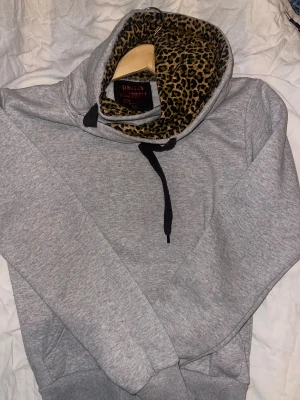Grå hoodie med leopard mönster xs - Säljer en grå hoodie från United Marshall College i storlek XS. Insidan av huvan har ett fett leopardmönstrat foder som ger extra edge. Tröjan har svarta snören, känguruficka och ribbade muddar. Perfekt för dig som gillar att sticka ut med detaljer.