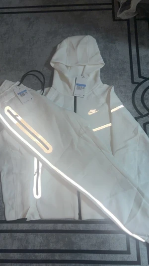 Nike vita mjukisbyxor med reflexdetaljer - Nike mjukisbyxor i vit färg med snygga reflexdetaljer längs benen och svarta dragkedjor. Byxorna har en modern, sportig look och är tillverkade i mjukt material som passar perfekt för chill eller träning. Snyggt Nike-logo på benet och smarta fickor med dragkedja.