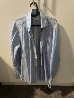 Blå randig skjorta från Stenströms - Snygg blå skjorta från Stenströms med smal passform och klassisk krage i storlek 39. Skjortan har vita och orangea ränder, ärmar med dubbla knappar vid manschetten och bröstficka. Tillverkad i exklusiv twofold super cotton för extra mjuk känsla. Perfekt för dig som gillar stilrena detaljer. Nypris:1800