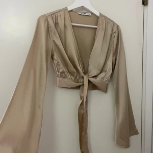 Champagnefärgad omlottopp från NA-KD - Säljer en champagnefärgad omlottopp från NA-KD i glansigt satinmaterial. Toppen har långa, vida ärmar och knyt framtill eller så kan man även snurra de runt midjan och knyta bak i ryggen istället! Båda sätt ger en snygg siluett. Croppad modell med djup v-ringning, perfekt för att lyfta din outfit.