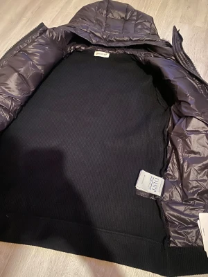 Svart pufferjacka från Moncler med huva - Svart pufferjacka från Moncler med stickade ärmar och huva. Jackan har quiltad framsida i glansig polyester och dragkedja hela vägen. Moncler-logga på ärmen och ribbad nederkant. Perfekt för dig som gillar stilren och sportig look. Nfc o tag funkar 