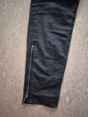 Jeansbyxor - Svarta skinny jeans från H&M med låg midja och ankellängd. Jeansen har snygga dragkedjor vid bensluten och klassisk femficksdesign. Materialet är stretchigt denim som sitter tight och följer kroppen. Perfekt för dig som gillar en smal siluett. 28 tum