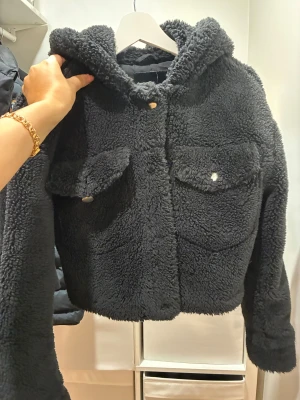 Svart teddyjacka från Pull&Bear - Mysig svart teddyjacka med huva från Pull&Bear. Jackan har två stora bröstfickor med knappar och en fluffig, mjuk yta som ger en cool och avslappnad vibe. Perfekt för kyliga dagar och enkel att matcha med jeans eller hoodies.