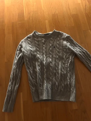 Grå kabelstickad tröja Polo Ralph Lauren - Snygg grå kabelstickad tröja från Polo Ralph Lauren i storlek XS. Tröjan har rund hals, långa ärmar och klassisk broderad logga på bröstet. Ribbstickade muddar vid ärmslut och nederkant ger en schysst passform. Perfekt för dig som gillar stilrena och tidlösa plagg.