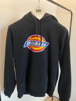 Svart Dickies hoodie i nyskick -storlek S - Svart hoodie från Dickies i storlek small med stor färgglad logga på bröstet. Tröjan har huva med snörning, magficka och långa ärmar. Perfekt för dig som gillar streetwear och vill ha en enkel men snygg look.