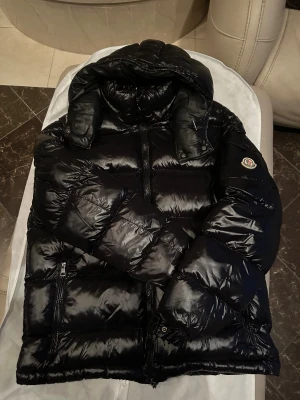 MONCLER MAYA Vinterjacka - Svart pufferjacka från Moncler med glansig finish och klassisk logga på ärmen. Jackan har huva, dragkedja framtill och quiltad design som ger en fet look. Perfekt för kalla dagar när du vill vara både varm och stilren. Jackan är i mycket gott skick som du kan se på bilderna. Vill du ha fler bilderna på skicket eller annat är det bara att fråga. Jackan är i storlek: 3(L)