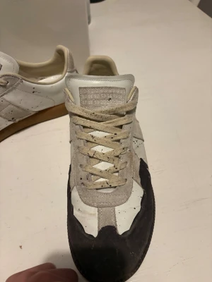Maison Margiela sneakers med färgstänk - Unika sneakers från Maison Margiela med vit och beige ovandel i skinn och mocka, bruna detaljer och coola svarta färgstänk. Klassisk rund tå, platt sula och snörning framtill. Perfekt för dig som gillar statement och designerstil. Hör av er vid minsta lilla fundering!!
