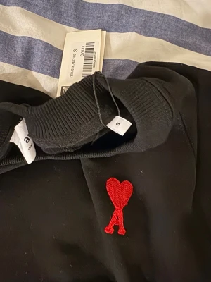 Svart sweatshirt från Ami Paris med rött hjärta - Svart sweatshirt från Ami Paris i storlek S med det ikoniska röda broderade hjärtat och bokstaven A på bröstet. Tröjan har rund hals, ribbade muddar och är långärmad. Perfekt för dig som gillar stilrena och trendiga plagg. S men passar M