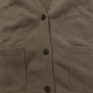 Beige stickad kofta med knappar - Mysig beige stickad kofta med ribbade muddar och två framfickor. Koftan har svarta knappar framtill och en klassisk v-ringad design. Perfekt för lager-på-lager och chill dagar. Materialet känns mjukt och värmande.