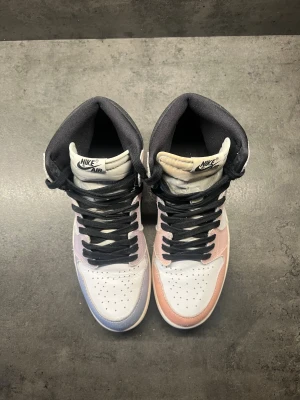 Nike Air Jordan 1 Pastell Highs - Säljer ett par Nike Air Jordan 1 Highs med unik pastellfärgning i rosa och lila toner, svart swoosh och svarta skosnören. Ovandelen är i textil och skinn, med klassisk rund tå och svart yttersula. Perfekta för dig som vill sticka ut med färg och stil.