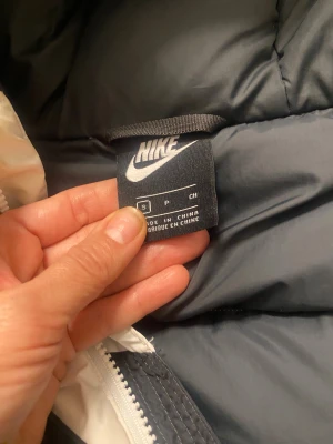 Svart och vit pufferjacka från Nike - Nike pufferjacka i svart och vitt med huva och feta dragkedjor. Jackan har en cool tvåfärgad design och logga på bröstet. Perfekt för kalla dagar när du vill ha både stil och värme. Snygg passform och flera fickor med dragkedja.