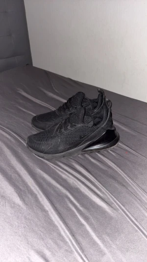 Svarta Nike Air Max 270 sneakers - Säljer ett par helsvarta Nike Air Max 270 sneakers med snygg mesh-överdel och stor synlig Air-enhet i hälen. Skorna har snörning, rund tå och platt sula. Perfekt för dig som gillar sportig och clean stil.