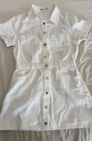 Vit jeansklänning från Zara, storlek L - Säljer en vit jeansklänning från Zara i storlek L. Köpte i 2024 men aldrig använt den. Klänningen har korta ärmar, krage, två bröstfickor och knappar hela vägen framtill. Den har även fickor på sidorna och baktill. 