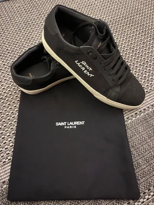 Svarta Saint Laurent sneakers - Svarta sneakers från Saint Laurent med vit logotyp på sidan. Skorna har snörning, rund tå och platt vit sula med fiskbensmönster undertill. Materialet är en mix av skinn och textil, vilket ger en clean och stilren vibe. Perfekta för dig som gillar exklusiva streetwear.