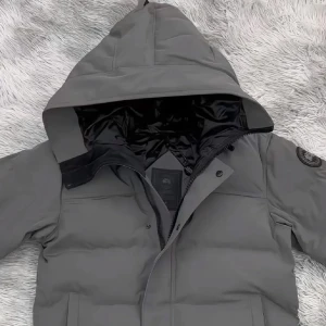 Grå dunjacka från Canada Goose - Säljer en grå dunjacka från Canada Goose med huva och svart logotyp på ärmen. Jackan har dragkedja och knappar framtill, två sidofickor och quiltad design. Perfekt för kalla dagar och riktigt snygg streetstil. Liten i str 