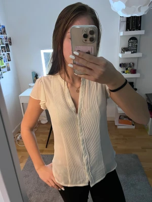 Vit blus med volangärm från H&M - Säljer en söt vit blus från H&M i storlek 36. Blusen har korta volangärmar, v-ringad hals och dekorativa veck framtill. Materialet är lätt och luftigt, perfekt för varmare dagar. Passformen är normal och den har fina detaljer längs knappraden.