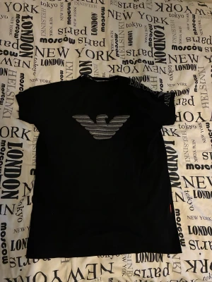 Svart Emporio Armani t-shirt M - Svart t-shirt från Emporio Armani i storlek M med klassisk rund hals och korta ärmar. Framtill finns det ikoniska Armani-logotypen i silver. Materialet är mjuk bomull som känns skönt mot huden. Perfekt för dig som gillar stilrena designer och exklusiva märken.