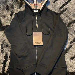 Svart Burberry hoodie med dragkedja - Svart hoodie från Burberry med klassisk rutmönstrad huva, broderad logga på bröstet och snörning. Tröjan har dragkedja framtill och ribbade muddar. Perfekt för dig som vill ha en stilren och lyxig look. Passar Xs och S 