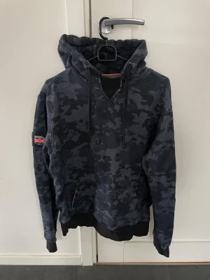 Mörkblå camo hoodie Superdry - Snygg mörkblå hoodie från Superdry med camouflagemönster i storlek M, modellen är ca 180cm. Hör av dig vid funderingar!