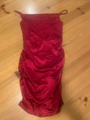 Röd sammetsklänning med smala axelband - Snygg röd kortklänning i mjuk sammet med smala axelband och rynkade detaljer längs sidorna. Klänningen har en figurnära passform och är perfekt för dig som vill sticka ut med en djärv färg och lyxig känsla. Passar dig som gillar trendiga och eleganta plagg.