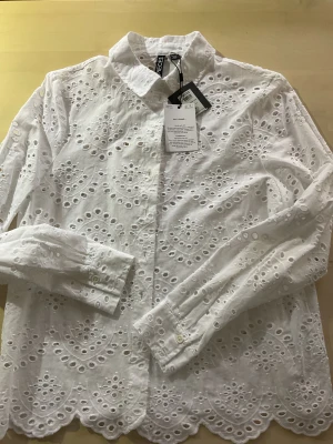 Vit broderad skjorta från Pieces - Supersnygg vit skjorta från Pieces i 100% bomull med broderade detaljer och hålmönster. Skjortan har klassisk krage, knappar framtill och långärmade ärmar. Den har en scallopad kant nedtill som ger en extra cool touch. Perfekt för dig som gillar unika plagg med mycket detaljer.